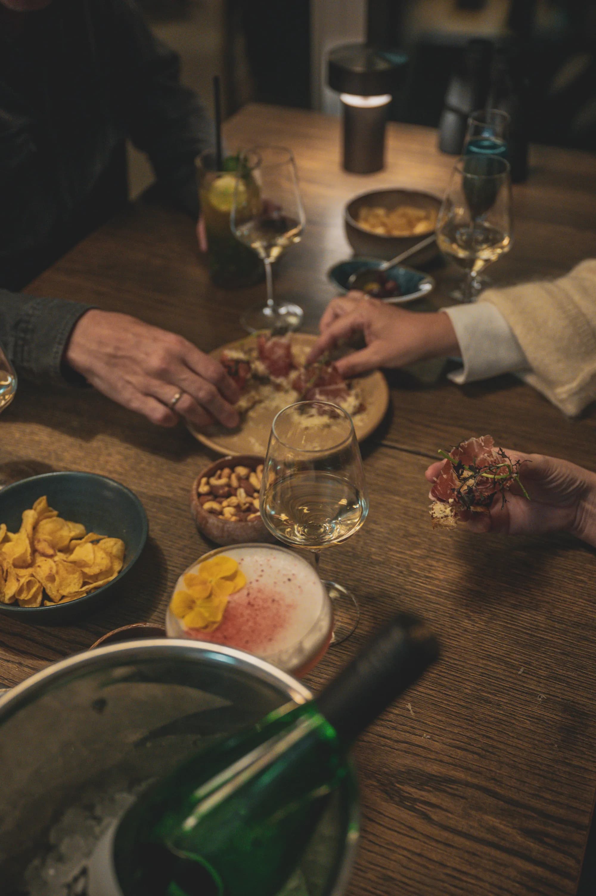 Hands reaching across a shared table of small plates and wine